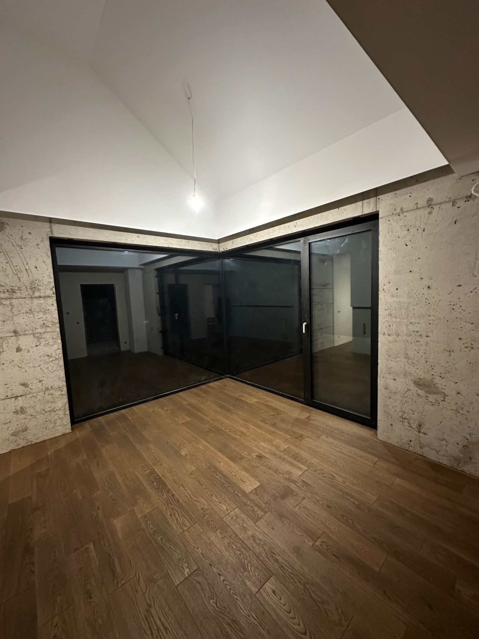 Nowoczesne wnętrze z drewnianą podłogą i szklanymi drzwiami przesuwnymi w czarnych ramach. Surowe betonowe ściany i biały sufit tworzą kontrastowe wykończenie.