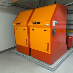 FABIOTECH Roman Pyrdoł - Dwa pomarańczowo-czerwone urządzenia grzewcze Hoval, stojące na podwyższonej betonowej platformie w pomieszczeniu technicznym, z widocznymi rurami i kanałami wentylacyjnymi.