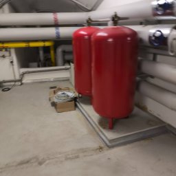 FABIOTECH Roman Pyrdoł - Dwa czerwone zbiorniki ciśnieniowe w kotłowni, otoczone białymi i żółtymi rurami z oznaczeniami. Na betonowej podłodze leży karton z kablami. Ujęcie z lekkim rozmyciem.