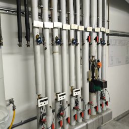FABIOTECH Roman Pyrdoł - System rur grzewczych lub chłodniczych z białą izolacją, zaworami, licznikami i pompą, zamontowany na białej ścianie w pomieszczeniu technicznym.
