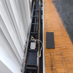 FABIOTECH Roman Pyrdoł - Otwarta instalacja ogrzewania kanałowego z izolowanymi rurami, mosiężnymi zaworami i grzejnikiem, obok białych żaluzji pionowych i drewnianej podłogi. Widoczna zdejmowana pokrywa.