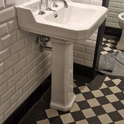 FABIOTECH Roman Pyrdoł - Klasyczna biała umywalka na postumencie z chromowaną baterią, na tle białych kafelków typu metro z czarną listwą i podłogą w czarno-białą szachownicę.