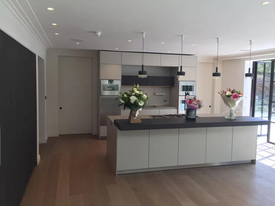 Nowoczesna kuchnia z wyspą, kwiatami i drewnianą podłogą. Jasne szafki, czarne blaty i wiszące lampy. Widok na ogród przez przeszklone drzwi.