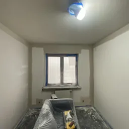 Pokój w trakcie przygotowania do malowania: zabezpieczone folią podłoga i meble, oklejone okno, widoczna lampa i narzędzia malarskie. Ściany częściowo zagruntowane.