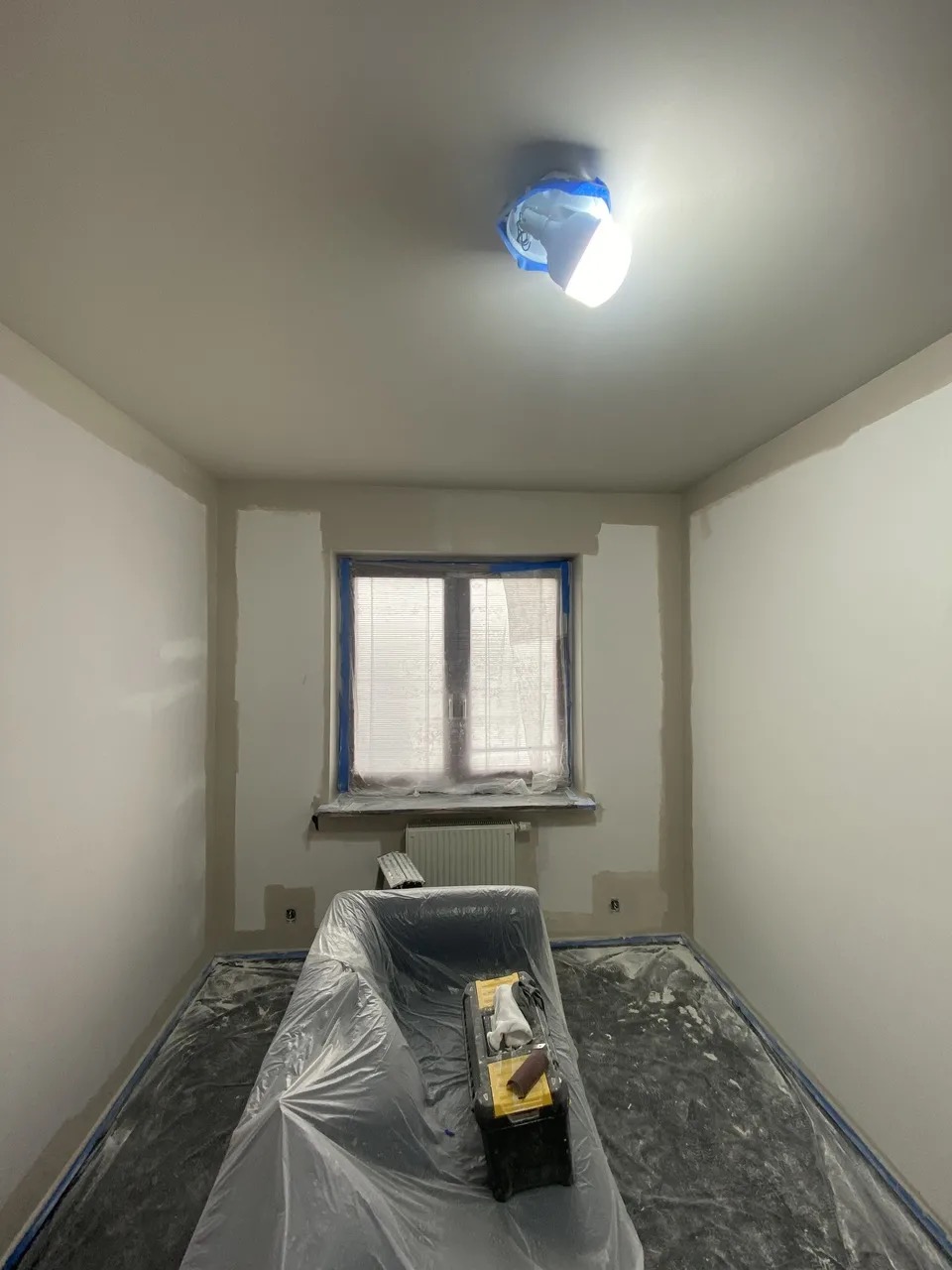 Pokój w trakcie przygotowania do malowania: zabezpieczone folią podłoga i meble, oklejone okno, widoczna lampa i narzędzia malarskie. Ściany częściowo zagruntowane.