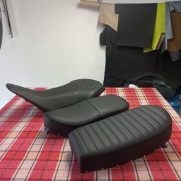 Tap1cer - Tapicerowane, czarne siedzenia motocyklowe z przeszyciami, ułożone na kraciastej tkaninie. W tle skóry tapicerskie i lampa warsztatowa.