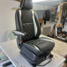 Odnowione, czarne, tapicerowane fotel kierowcy z regulowanym zagłówkiem i podłokietnikiem, z białymi wstawkami i romboidalnym przeszyciem, na warsztatowym stole.