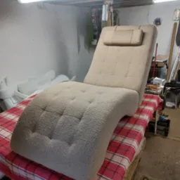 Tapicerowany leżak w beżowym odcieniu z zagłówkiem, umieszczony na stole z czerwoną kratą w warsztacie. Widoczne narzędzia i materiały tapicerskie w tle.