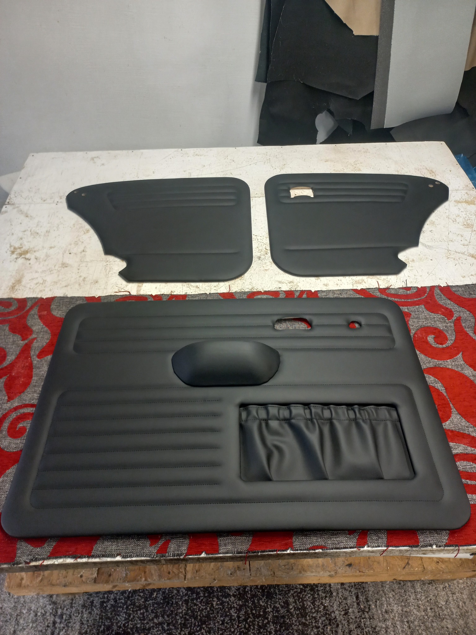 Odnowione, czarne panele drzwiowe samochodu na stole tapicerskim, z widocznymi detalami przeszyć i kieszenią. Tkaniny w tle sugerują warsztat.