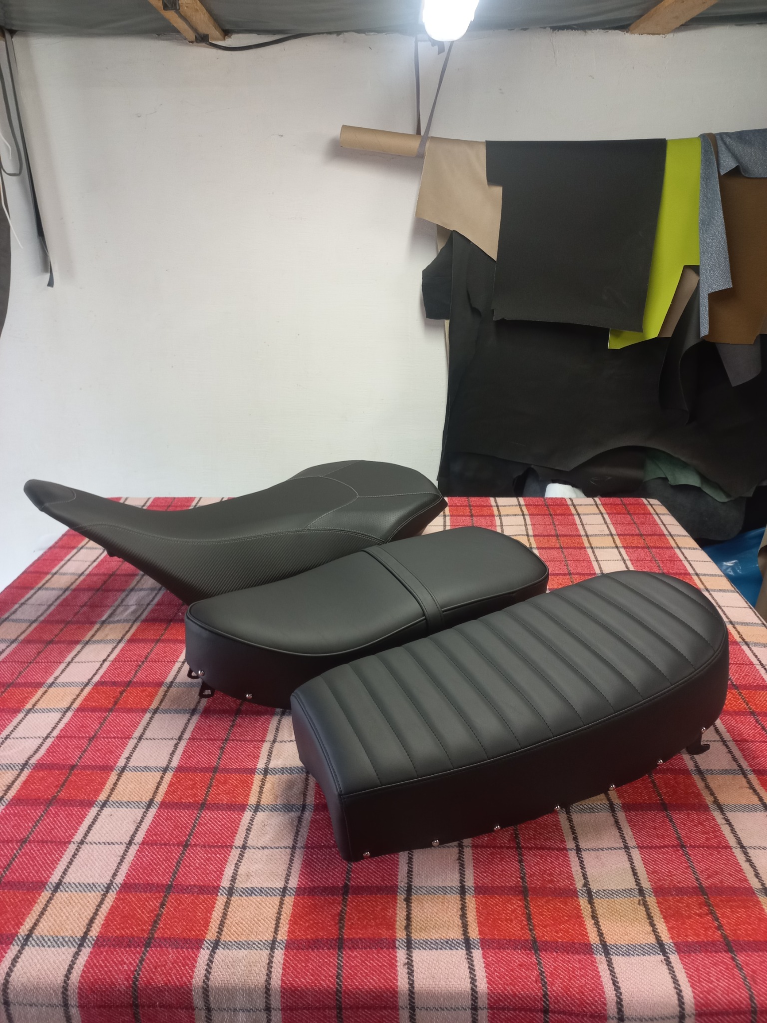 Tapicerowane, czarne siedzenia motocyklowe z przeszyciami, ułożone na kraciastej tkaninie. W tle skóry tapicerskie i lampa warsztatowa.