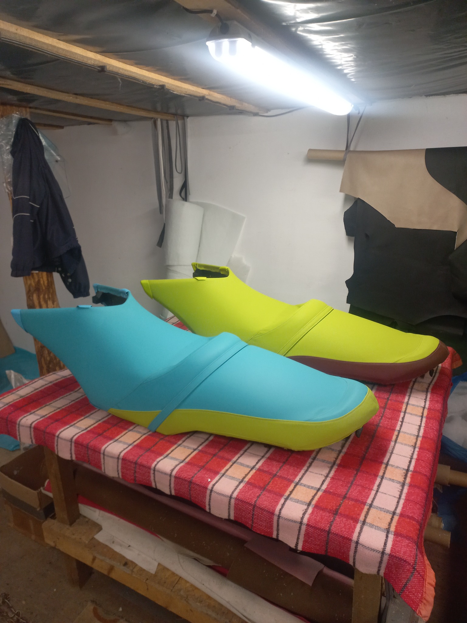 Dwa jaskrawo kolorowe, tapicerowane elementy siedzisk na stole w warsztacie, z widocznymi kawałkami skóry i narzędziami w tle.