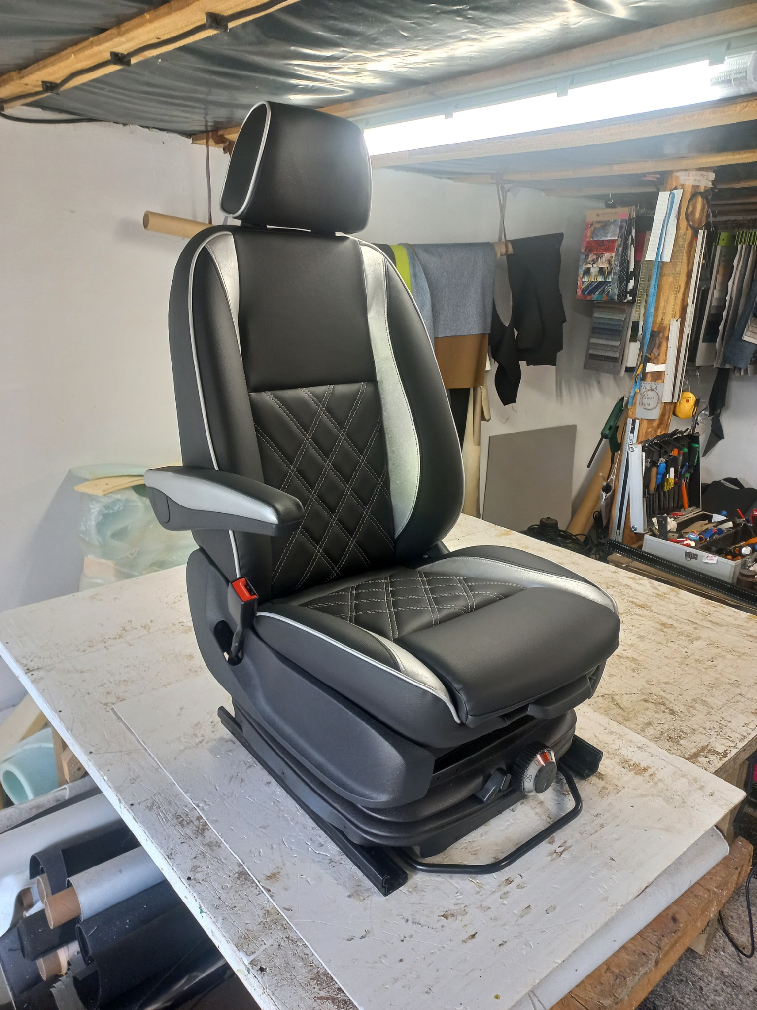 Odnowione, czarne, tapicerowane fotel kierowcy z regulowanym zagłówkiem i podłokietnikiem, z białymi wstawkami i romboidalnym przeszyciem, na warsztatowym stole.