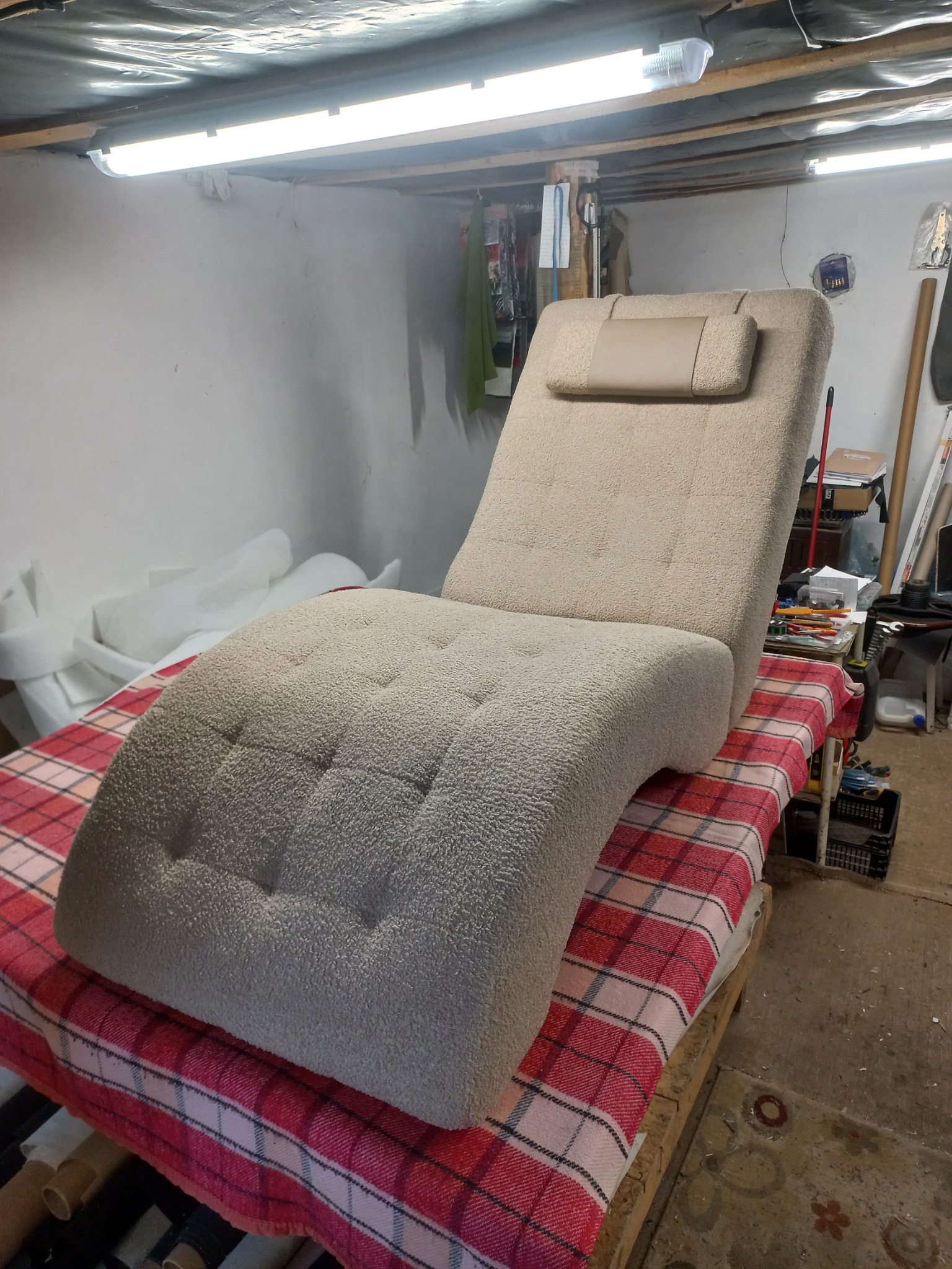 Tapicerowany leżak w beżowym odcieniu z zagłówkiem, umieszczony na stole z czerwoną kratą w warsztacie. Widoczne narzędzia i materiały tapicerskie w tle.