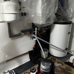 Grzegorz Rotter - Biały zbiornik wyrównawczy i bojler w folii ochronnej, szare rury, pompa do wody i skrzynka narzędziowa na dywanie. Instalacja w trakcie prac.