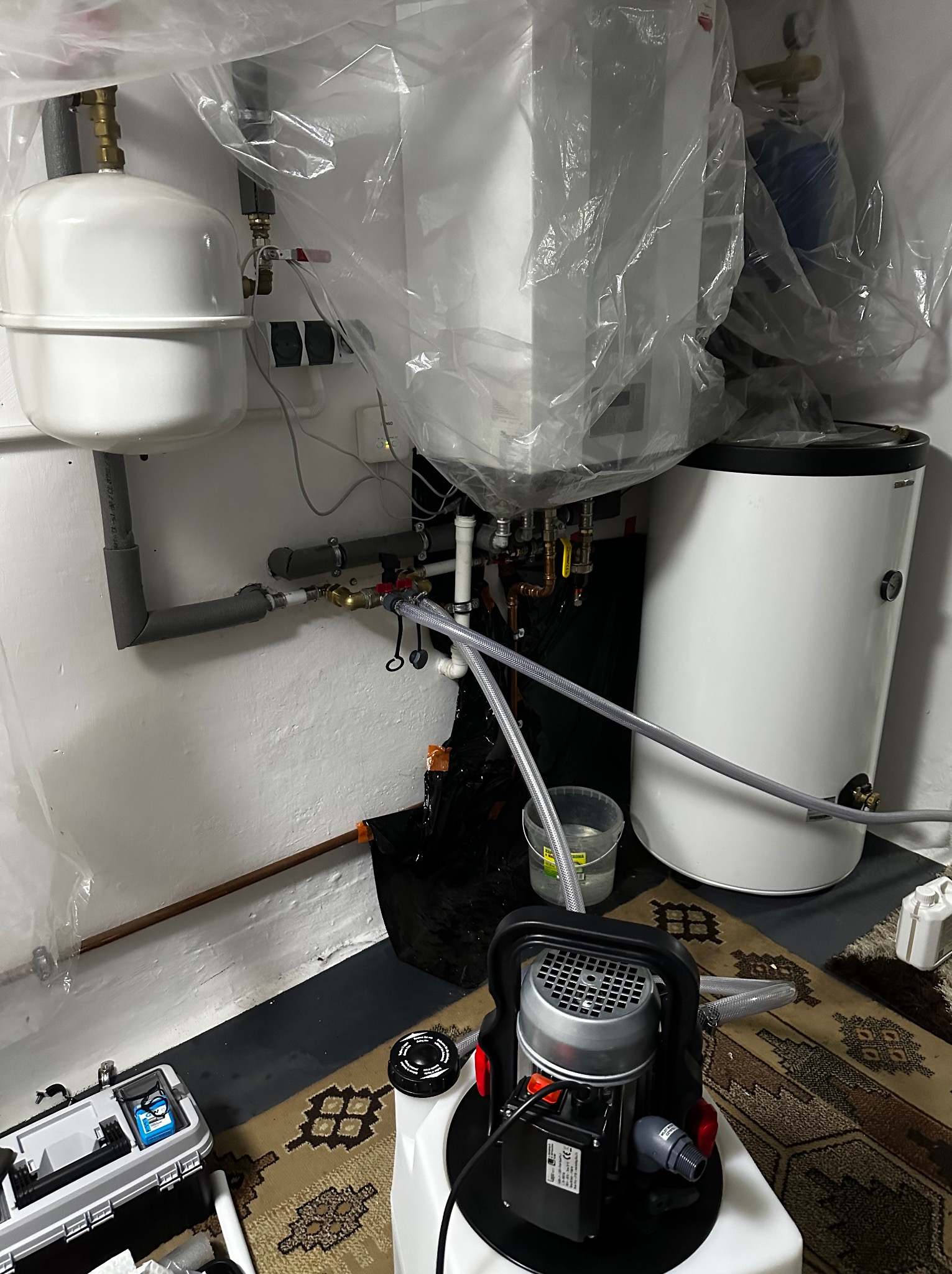 Biały zbiornik wyrównawczy i bojler w folii ochronnej, szare rury, pompa do wody i skrzynka narzędziowa na dywanie. Instalacja w trakcie prac.