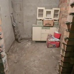 Zdemontowane meble i sprzęty w piwnicy, ceglane fragmenty ścian, ślady po remoncie, drabina oparta o ścianę, betonowa podłoga. Przygotowanie do opróżniania.