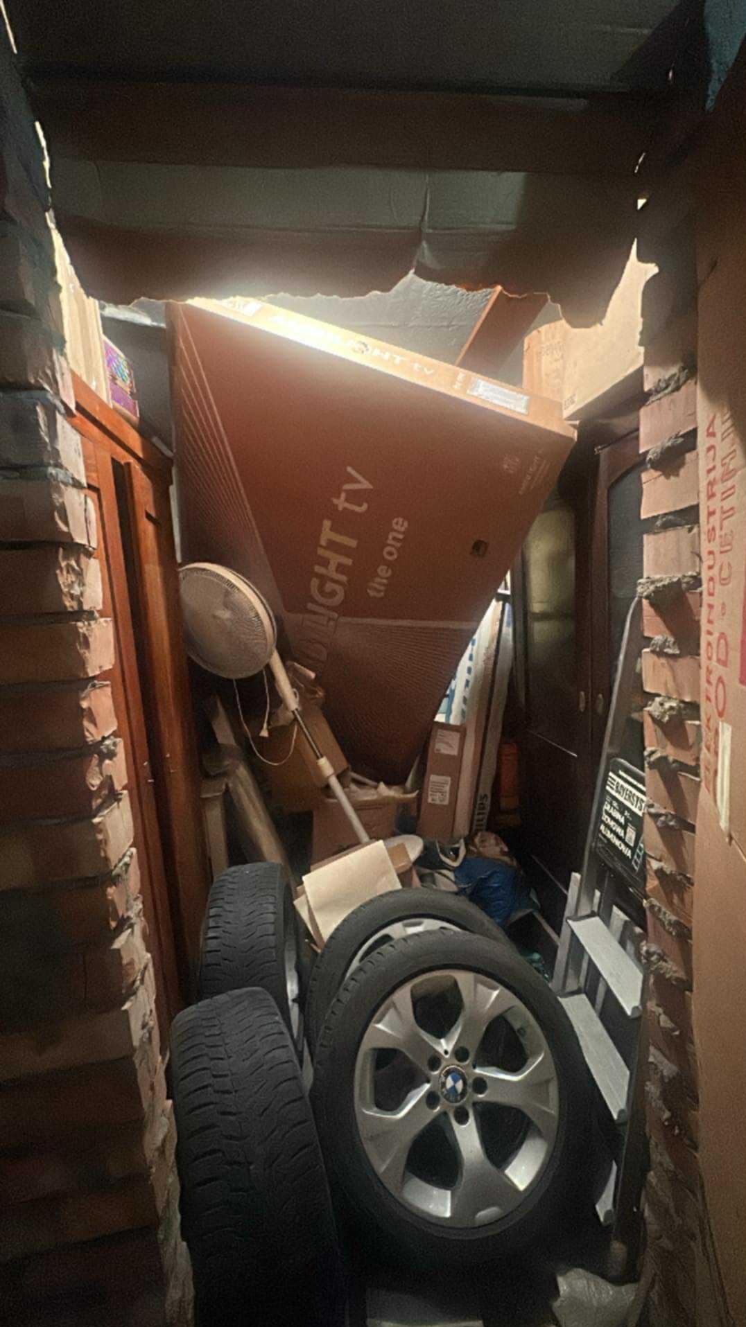 Zastawiony korytarz z kartonami, oponami i sprzętem AGD. Widoczne ceglane ściany i schody. Chaos przestrzeni sugeruje potrzebę opróżnienia.