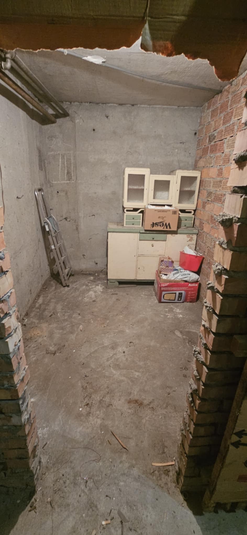 Zdemontowane meble i sprzęty w piwnicy, ceglane fragmenty ścian, ślady po remoncie, drabina oparta o ścianę, betonowa podłoga. Przygotowanie do opróżniania.