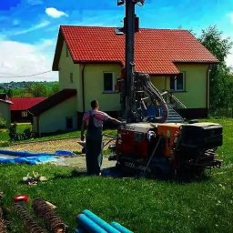 ZAINSTAL Usługi wodno-kanalizacyjne, wykończeniowe Małgorzata Pręgowska - Prace wiertnicze na posesji: operator obsługuje maszynę wiertniczą na tle domu z czerwoną dachówką. Widoczne niebieskie rury i rozłożona folia. Słoneczny dzień.