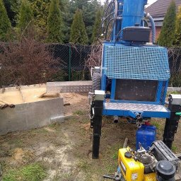 ZAINSTAL Usługi wodno-kanalizacyjne, wykończeniowe Małgorzata Pręgowska - Niebieska wiertnica do studni w trakcie pracy. Widoczny pracownik, rury, zbiornik na wodę i błoto. Tło stanowią drzewa i ogrodzenie.