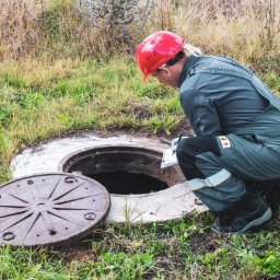 ZAINSTAL Usługi wodno-kanalizacyjne, wykończeniowe Małgorzata Pręgowska - Pracownik w czerwonym kasku, w kombinezonie, notuje dane przy otwartym włazie studzienki kanalizacyjnej na zewnątrz, w otoczeniu trawy i krzewów.