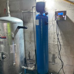 ZAINSTAL Usługi wodno-kanalizacyjne, wykończeniowe Małgorzata Pręgowska - Hydrofor ze zbiornikiem ocynkowanym i niebieski filtr wody z elektronicznym sterowaniem, zainstalowane w pomieszczeniu z surowymi ścianami.