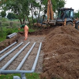 ZAINSTAL Usługi wodno-kanalizacyjne, wykończeniowe Małgorzata Pręgowska - Budowa drenażu: pracownik układa rury drenarskie w wykopie wypełnionym żwirem, w tle koparka i hałda ziemi. Widoczne elementy instalacji i krajobraz wiejski.