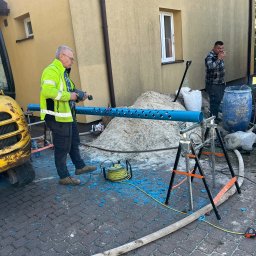 ZAINSTAL Usługi wodno-kanalizacyjne, wykończeniowe Małgorzata Pręgowska - Pracownik w żółtej kamizelce wierci otwory w niebieskiej rurze na tle budynku z numerem 25. Widoczny sprzęt budowlany i drugi pracownik w tle.