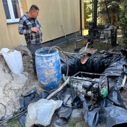 ZAINSTAL Usługi wodno-kanalizacyjne, wykończeniowe Małgorzata Pręgowska - Wiercenie studni: mężczyzna w kraciastej koszuli nadzoruje proces wiercenia, widoczna wiertnica, pompa, niebieska beczka i wykop zabezpieczony folią.