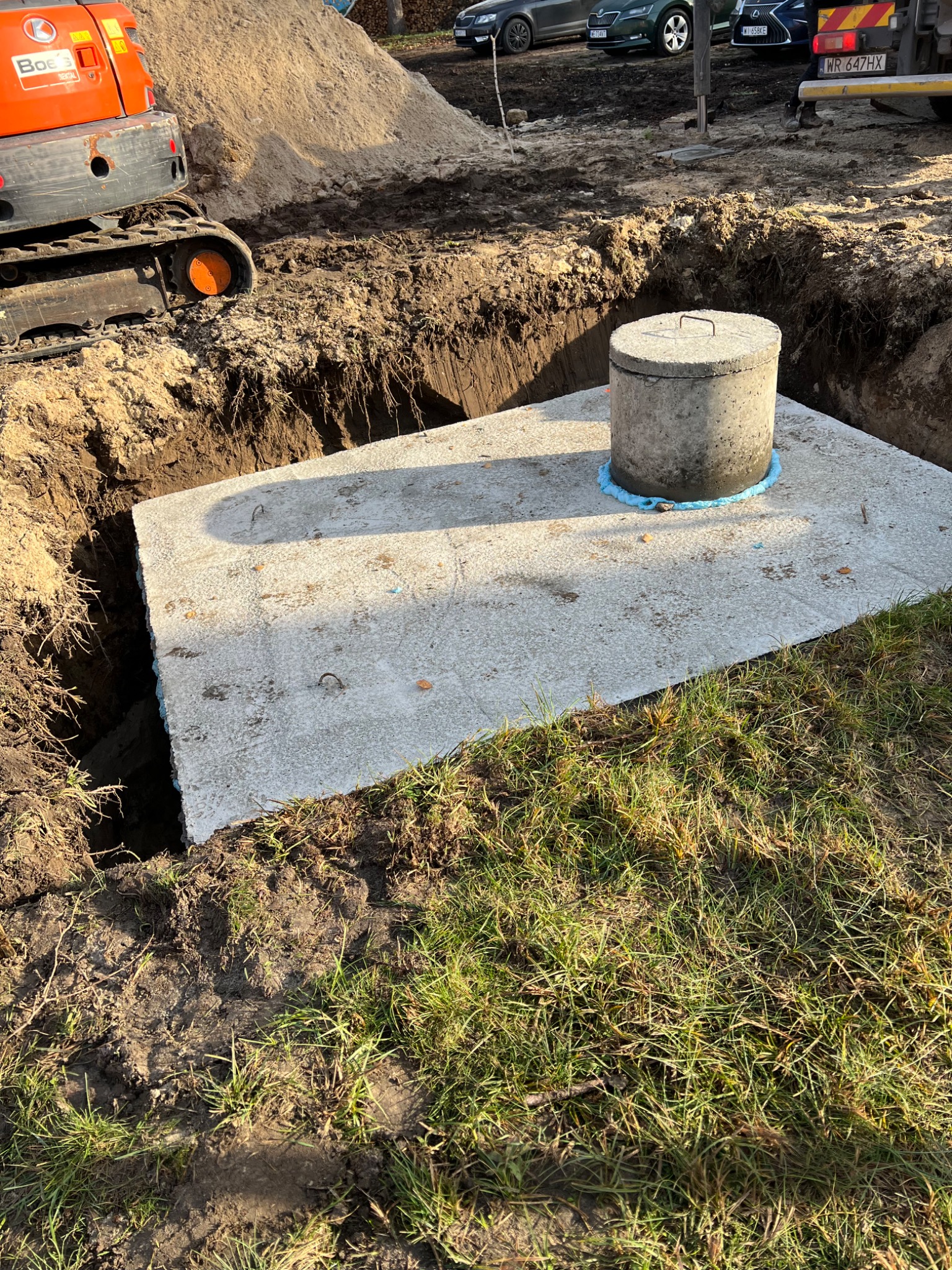 Betonowy zbiornik z włazem w wykopie, obok minikoparka. Widoczne ślady ziemi i trawy. Słoneczny dzień, częściowo zacienione ujęcie. Prace ziemne.