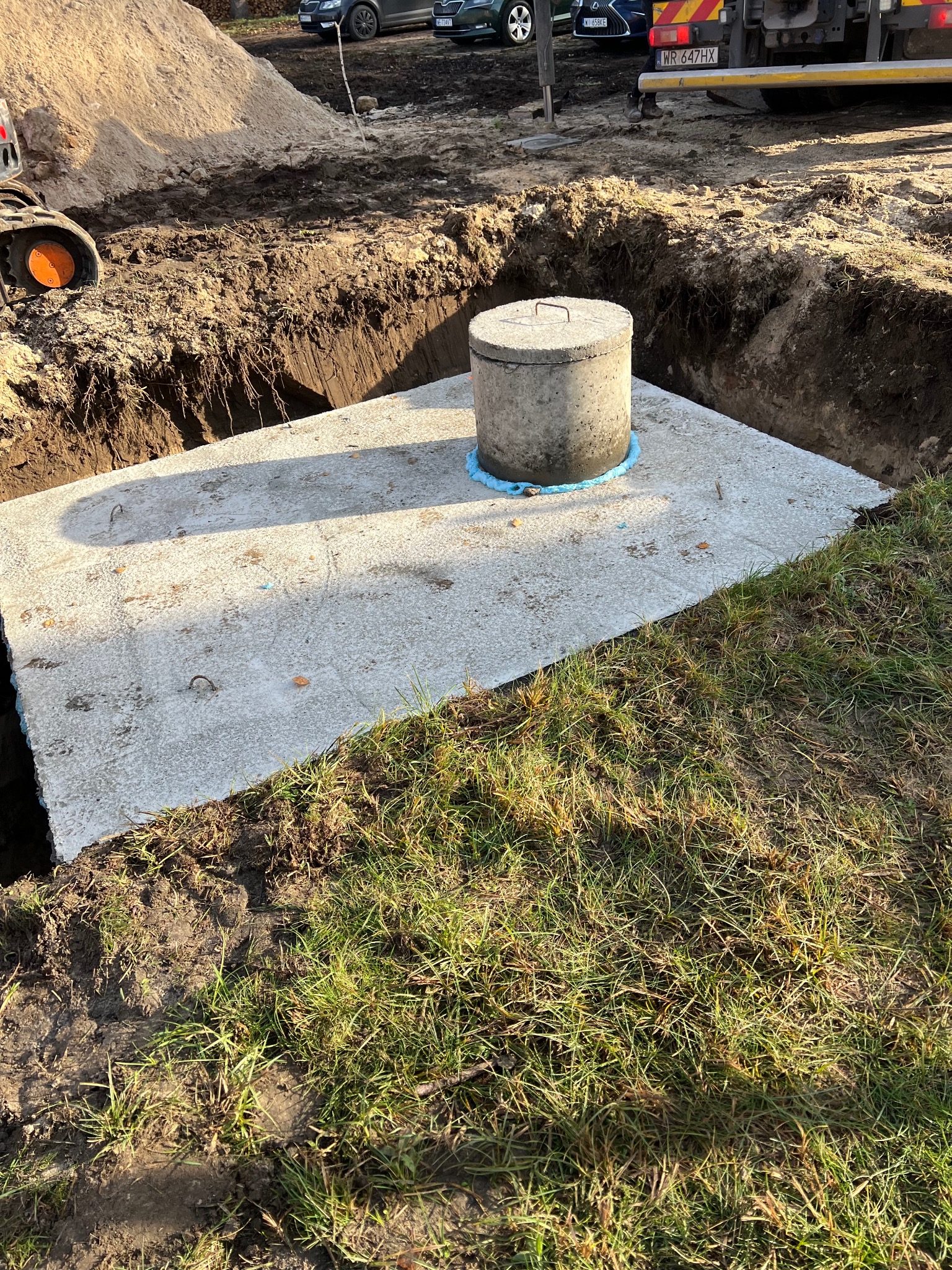 Betonowy zbiornik z pokrywą, umieszczony w wykopie ziemnym, otoczony trawą. Widoczny fragment koparki i zaparkowane samochody w tle. Słoneczny dzień.