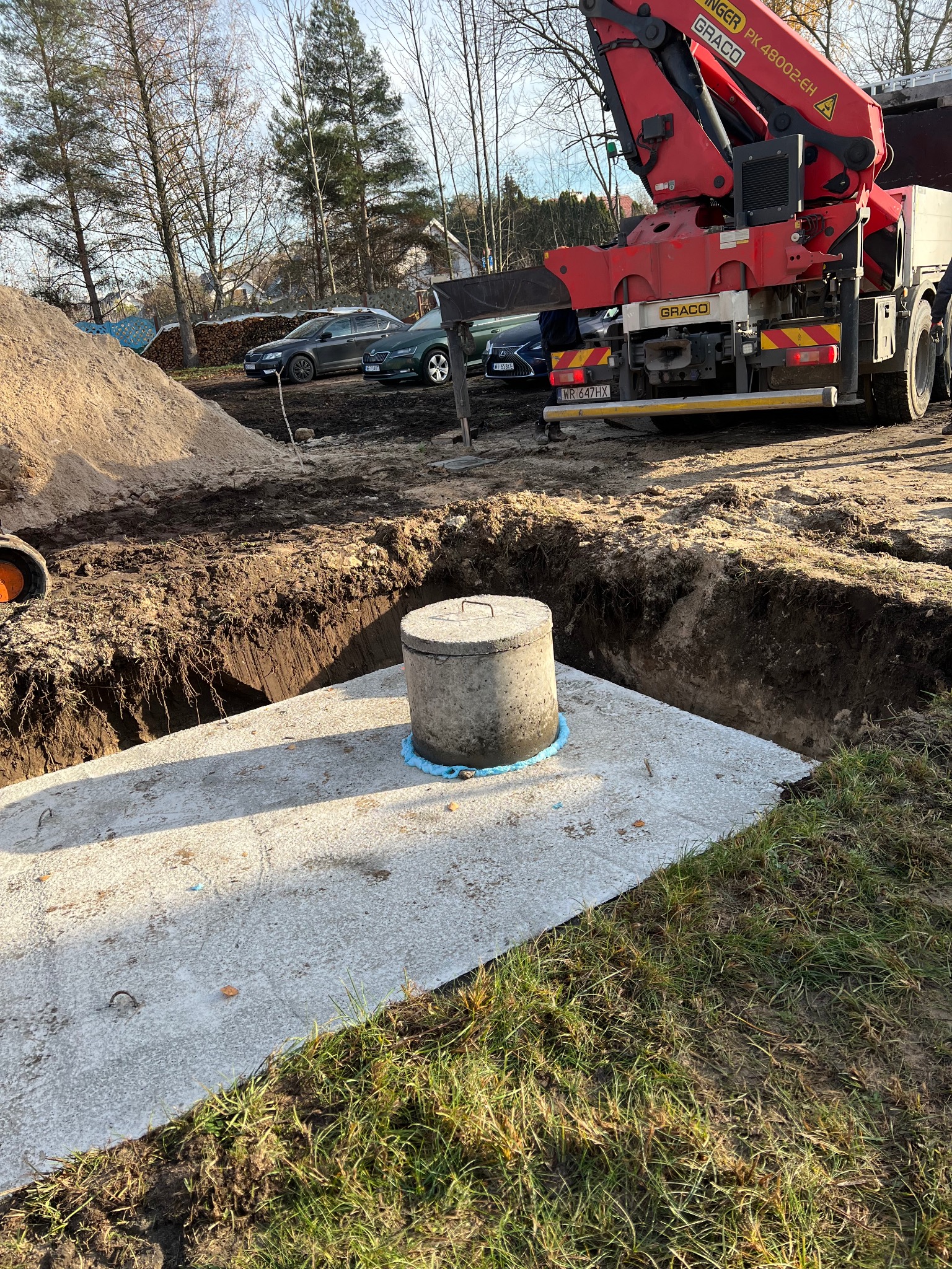 Betonowy zbiornik w wykopie, zabezpieczony niebieską taśmą, z dźwigiem w tle. Widoczny stos piasku i samochody. Słoneczny dzień.