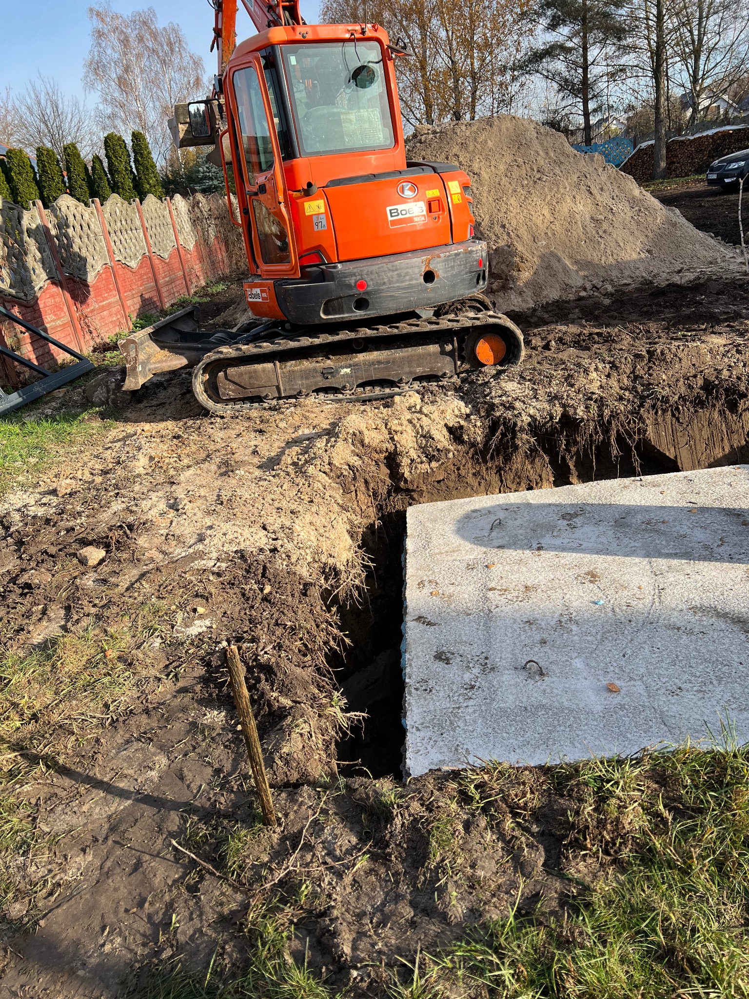 Pomarańczowa mini koparka obok wykopu z betonowym elementem. Stos ziemi w tle. Widok z góry na plac budowy w słoneczny dzień.