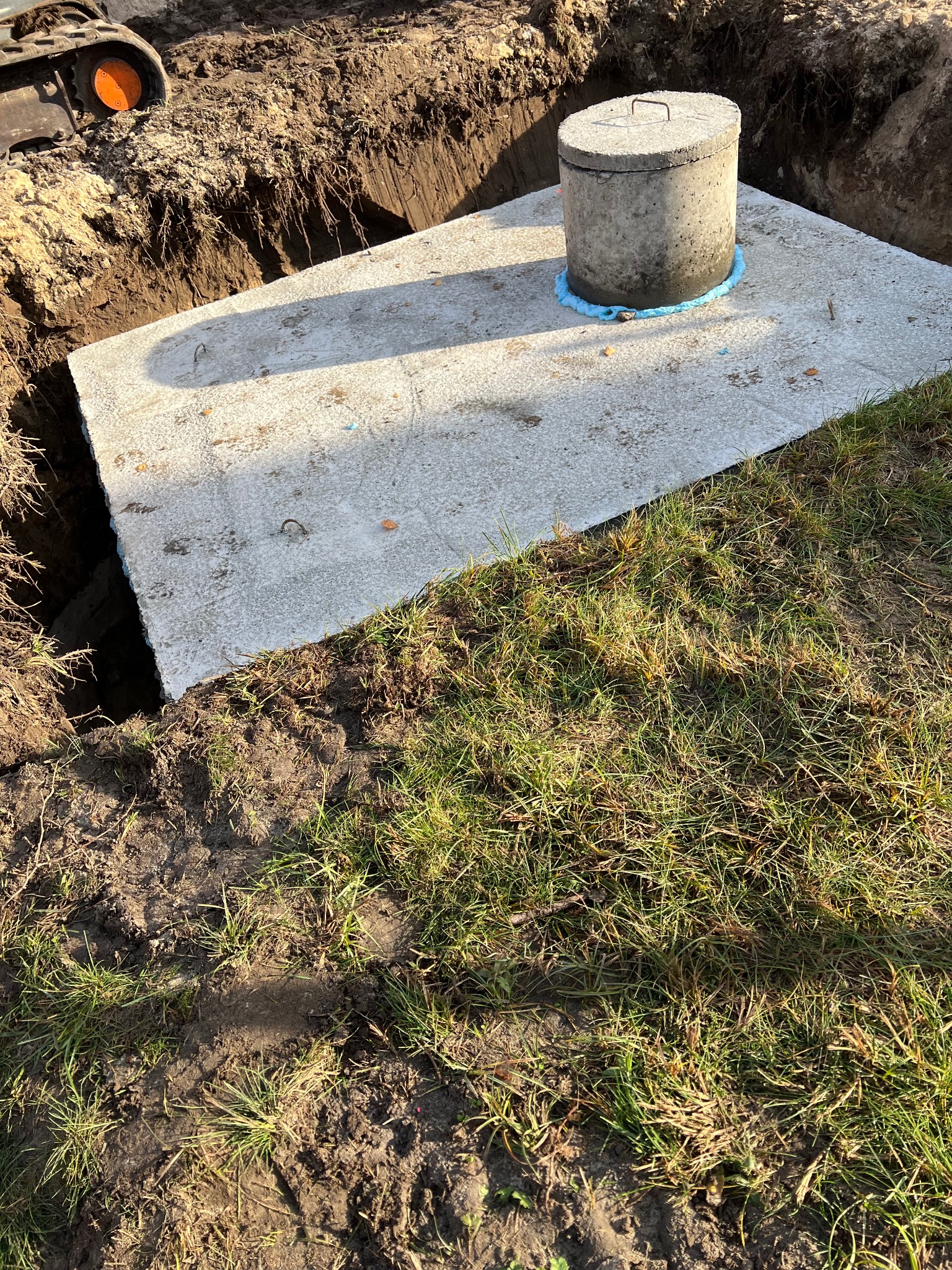 Betonowy zbiornik z pokrywą, częściowo zagłębiony w ziemi, z widocznym wykopem i fragmentem maszyny budowlanej w tle. Uszczelnienie wokół włazu.