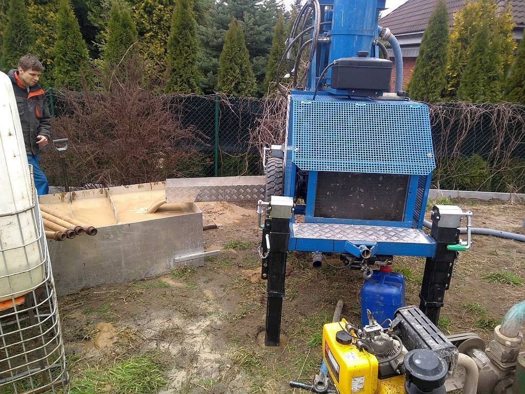 Niebieska wiertnica do studni w trakcie pracy. Widoczny pracownik, rury, zbiornik na wodę i błoto. Tło stanowią drzewa i ogrodzenie.