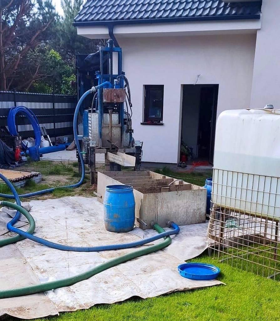 Wiertnica studni głębinowej na posesji, z widocznymi elementami instalacji: zbiornik na wodę, beczka, węże. Prace w toku, teren zabezpieczony folią. Dom z ciemną dachówką w tle.