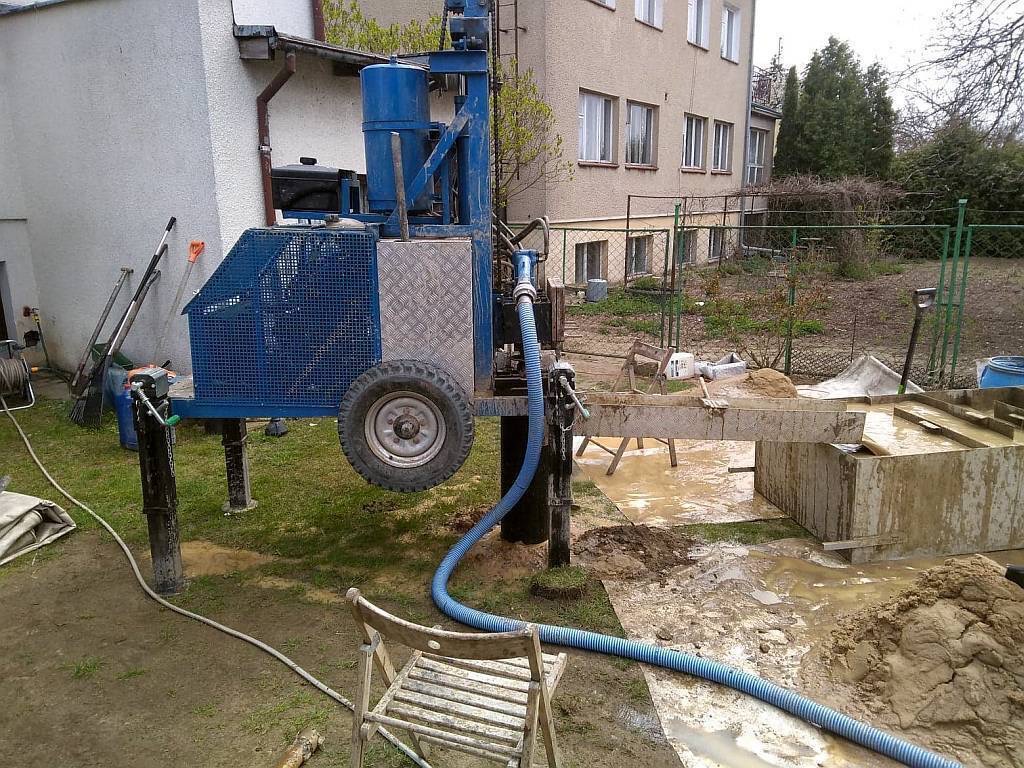 Niebieska wiertnica mobilna na trawniku, z widocznym osprzętem i błotem. W tle budynek mieszkalny i ogrodzenie. Ujęcie z bliska, ukazujące detale maszyny i otoczenia.