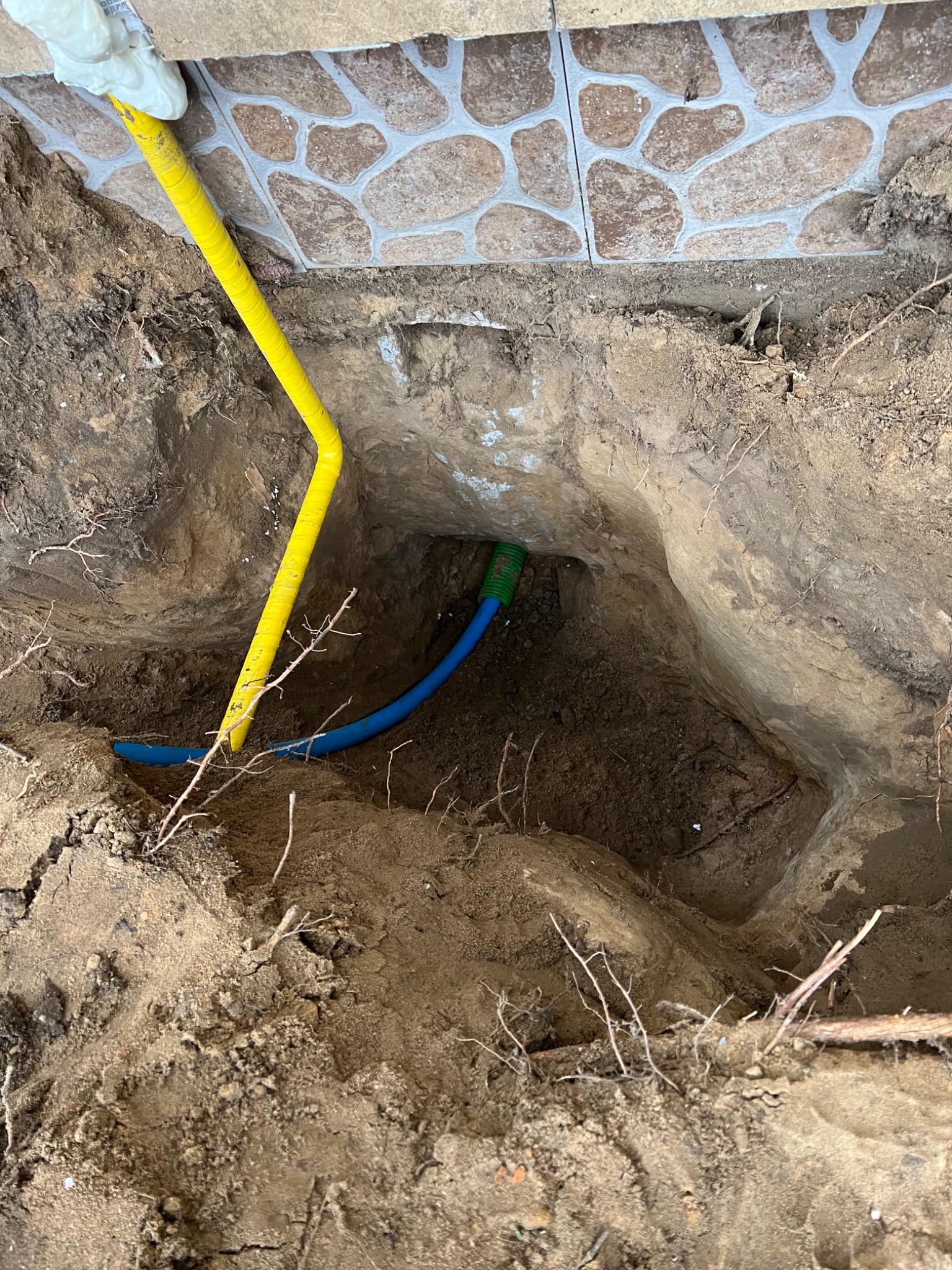 Żółta i niebieska rura w wykopie ziemnym przy fundamencie z kamiennej okładziny. Widoczne korzenie i fragment rękawicy ochronnej.