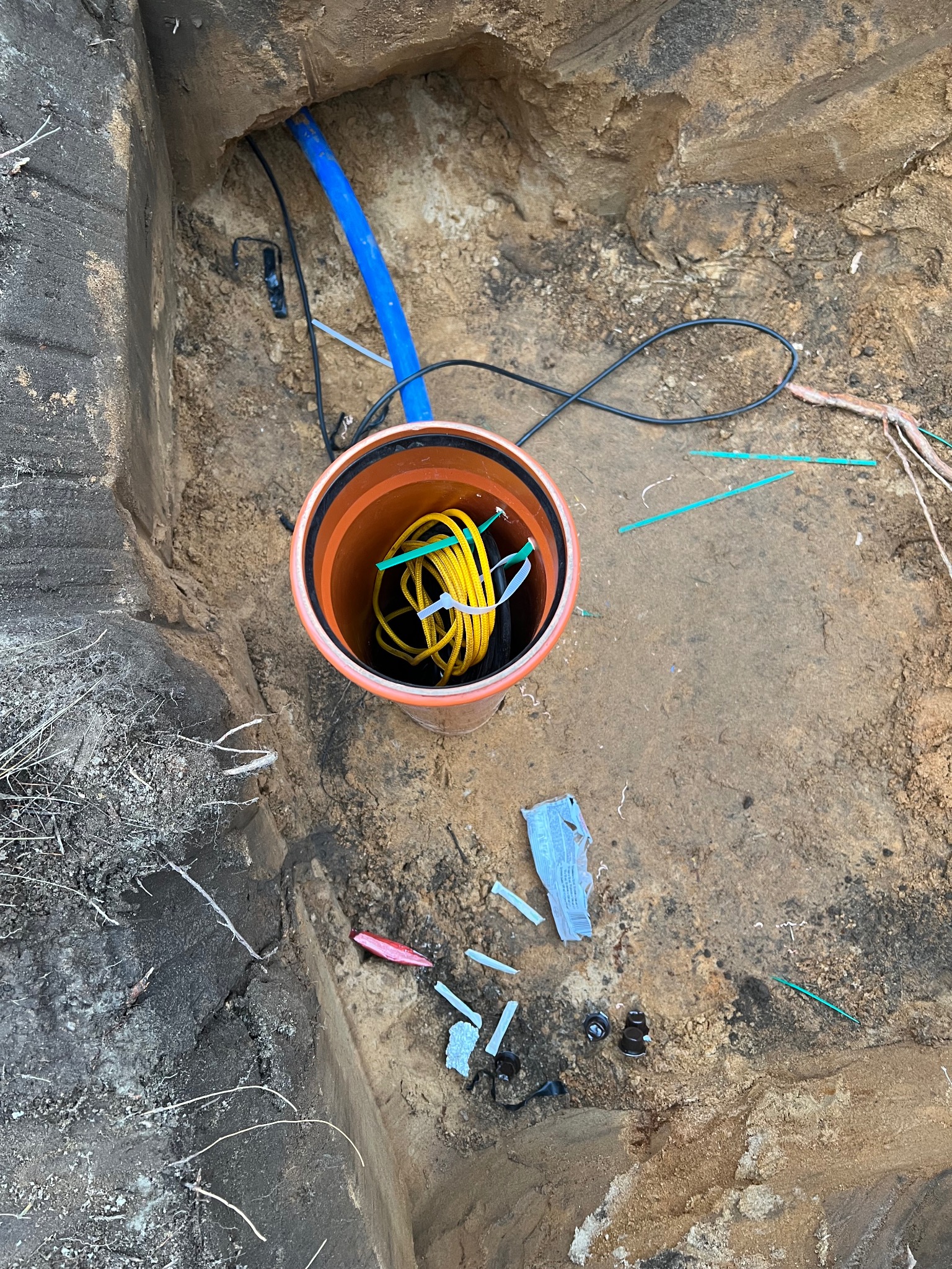 Wykop z rurą osłonową i kablem w ziemi. Niebieska rura wystaje z boku. Widoczne elementy instalacji i ziemia. Ujęcie z góry, podkreślające detale.