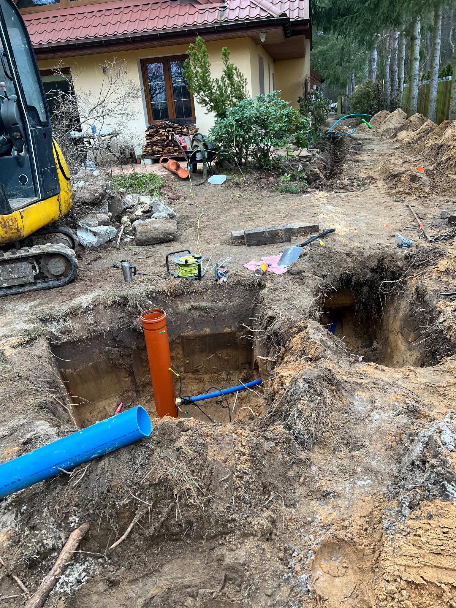 Wykop pod instalacje sanitarne z widocznymi rurami (pomarańczowa i niebieska) oraz fragmentem koparki. Prace ziemne w toku, teren wokół wykopu rozkopany.