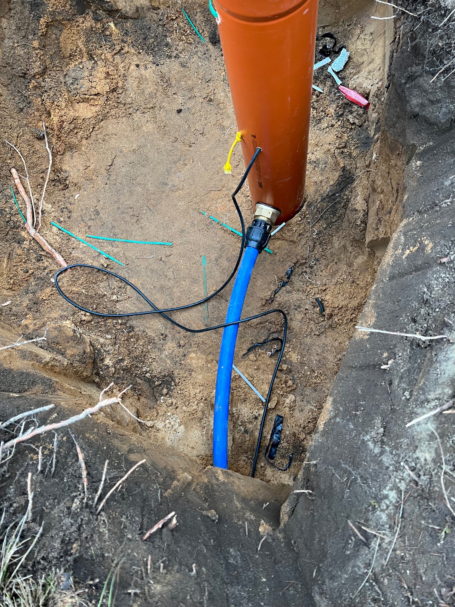 Brązowa rura w wykopie, połączona z niebieskim przewodem. Widoczne kable i elementy instalacji. Wykop ziemny z resztkami roślinności. Ujęcie z góry.