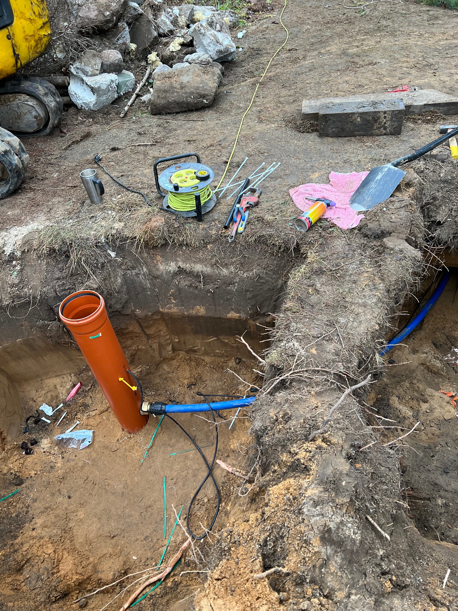 Wykop z pomarańczową rurą i niebieskim przewodem. Widoczne narzędzia, kabel zasilający i fragment koparki. Prace ziemne w trakcie realizacji.