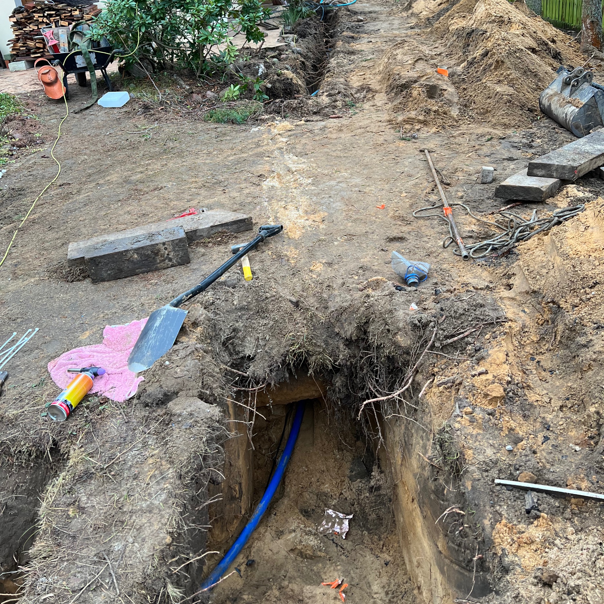 Wykopany rów z niebieską rurą w ziemi, widoczne narzędzia i sprzęt budowlany. Wykop pod instalacje w trakcie realizacji, ziemia, piasek i roślinność w tle.