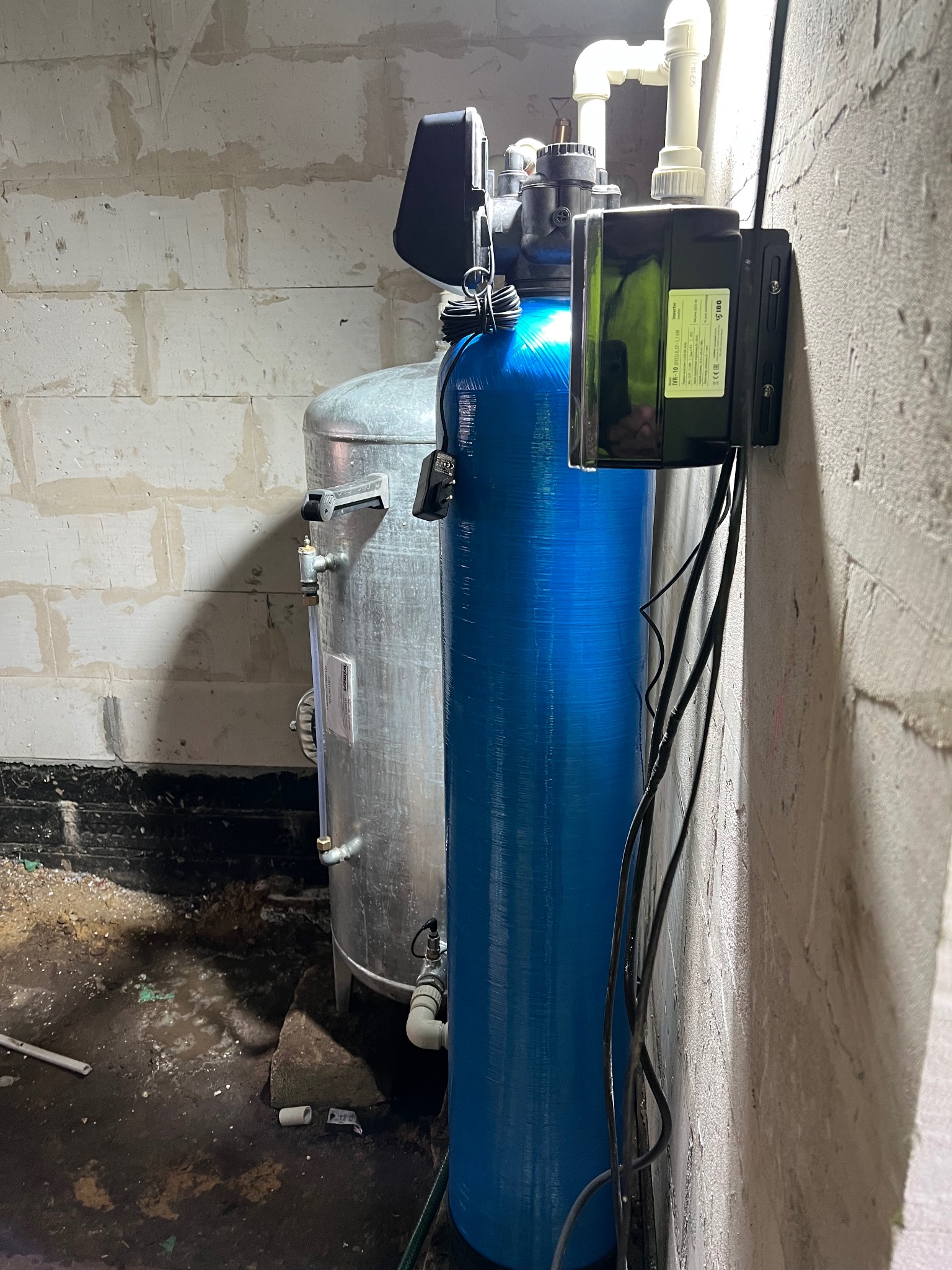 Niebieski i srebrny zbiornik hydroforowy z czarnym sterownikiem na ścianie z betonu komórkowego. Widoczne rury i przewody. Instalacja w piwnicy.