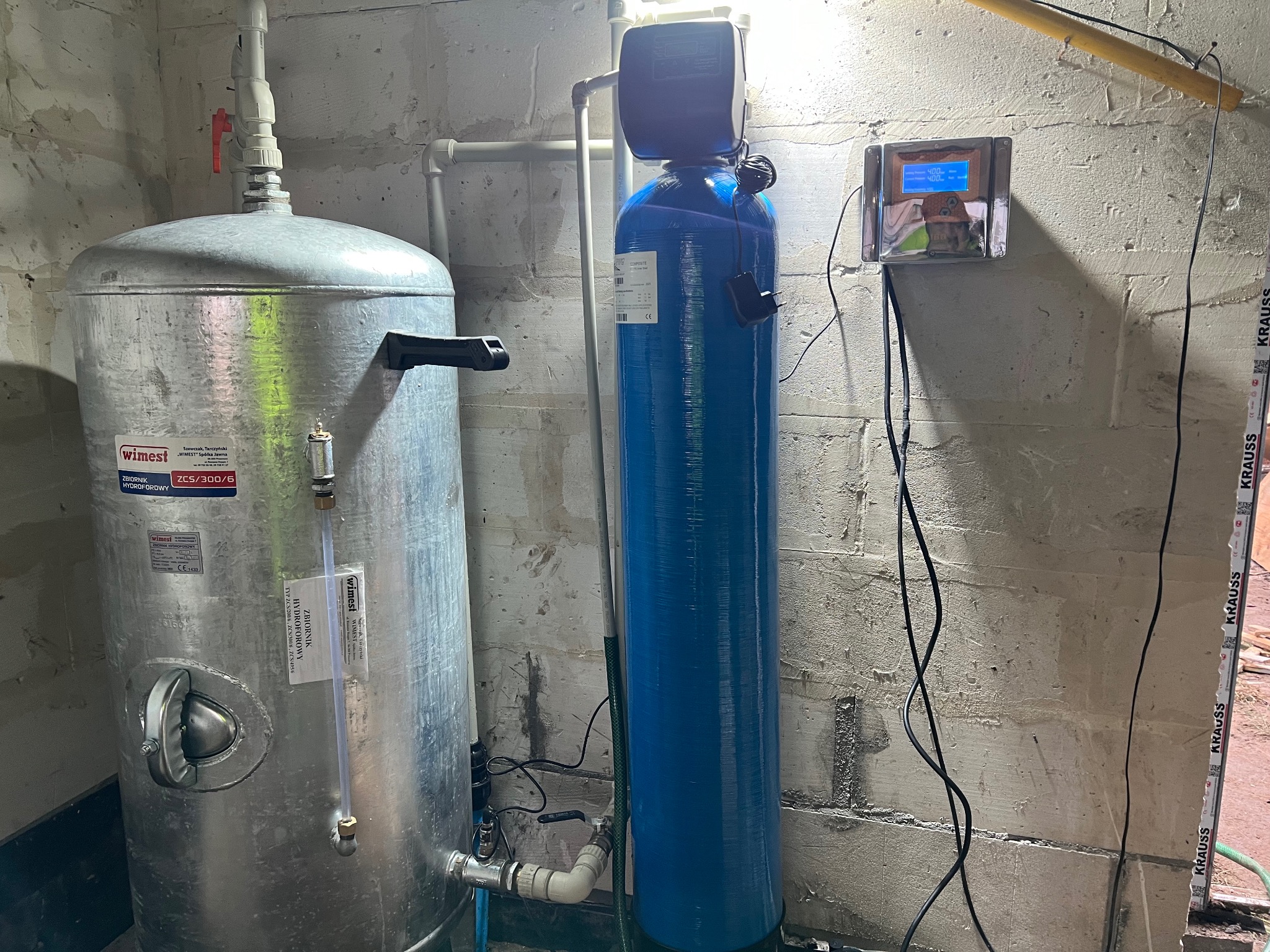 Hydrofor ze zbiornikiem ocynkowanym i niebieski filtr wody z elektronicznym sterowaniem, zainstalowane w pomieszczeniu z surowymi ścianami.