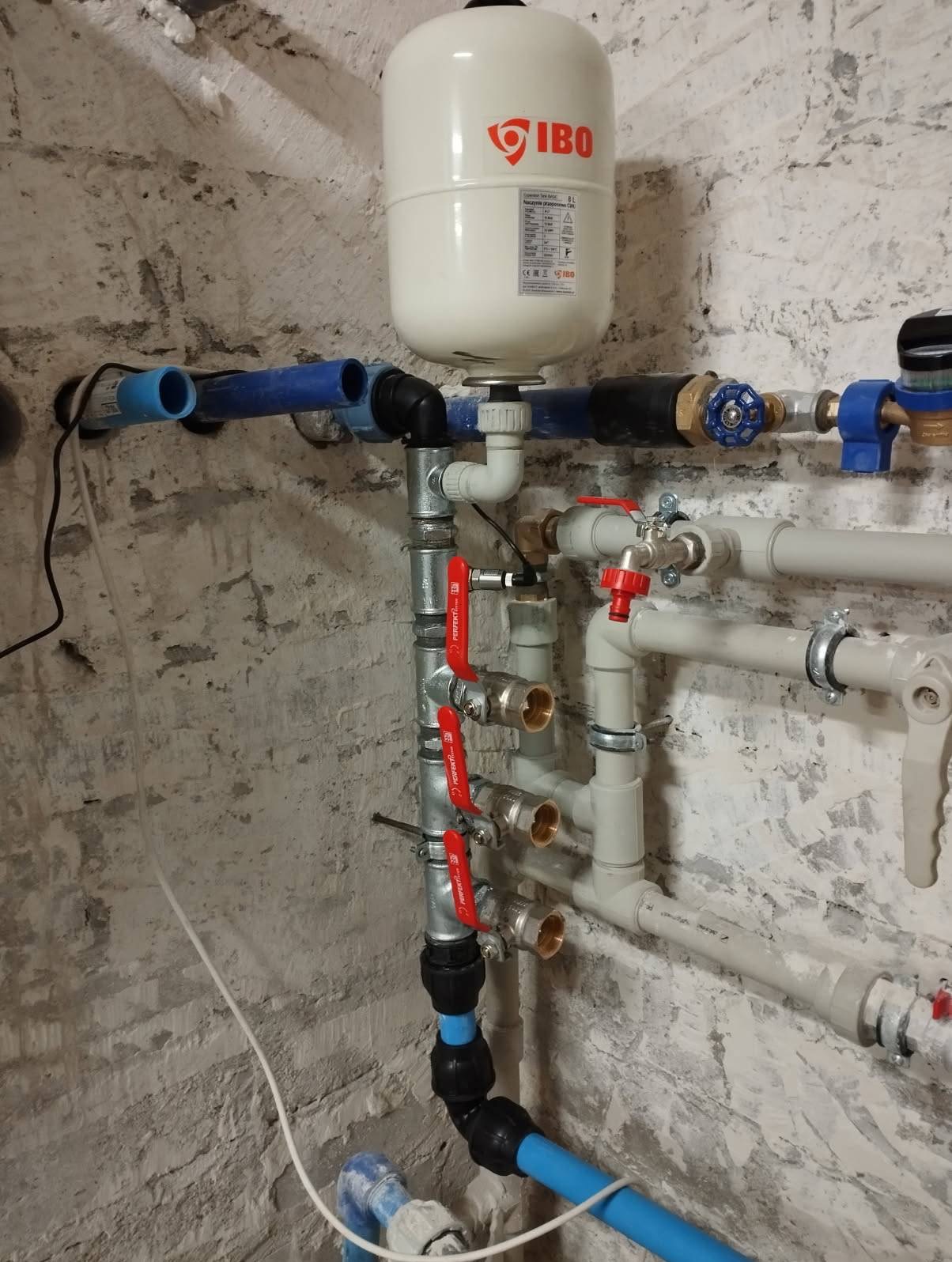 Złożona instalacja hydrauliczna z zbiornikiem IBO na tle surowej, białej ściany z cegły. Widoczne liczne rury, zawory z czerwonymi uchwytami i złączki.