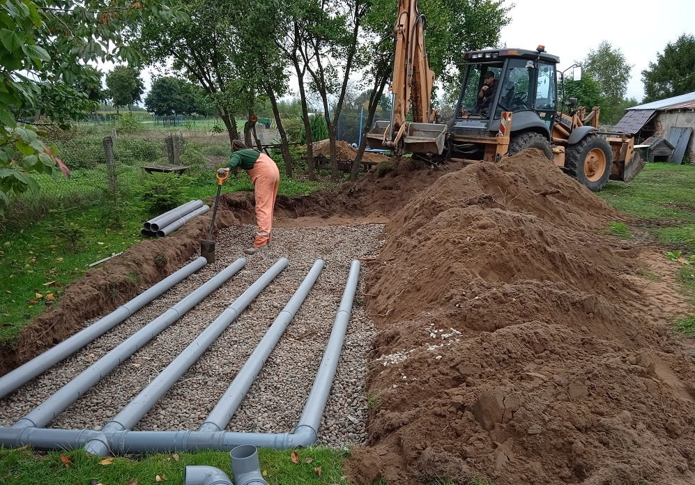 Budowa drenażu: pracownik układa rury drenarskie w wykopie wypełnionym żwirem, w tle koparka i hałda ziemi. Widoczne elementy instalacji i krajobraz wiejski.