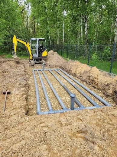 Instalacja drenażowa z rur ułożonych w wykopie, zasypanych żwirem, z minikoparką w tle, na tle zieleni. Widok z góry.