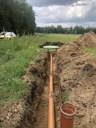 Brązowa rura w wykopie, zielona pokrywa studzienki w tle, trawa i drzewa. Instalacja kanalizacyjna na działce, widok z góry.