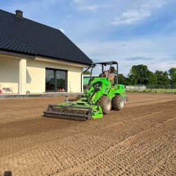 Luxform Garden Lutostańscy - Zielony traktor z wałem ugniata ziemię na wyrównanym terenie przed domem. Przygotowanie pod trawnik w słoneczny dzień.